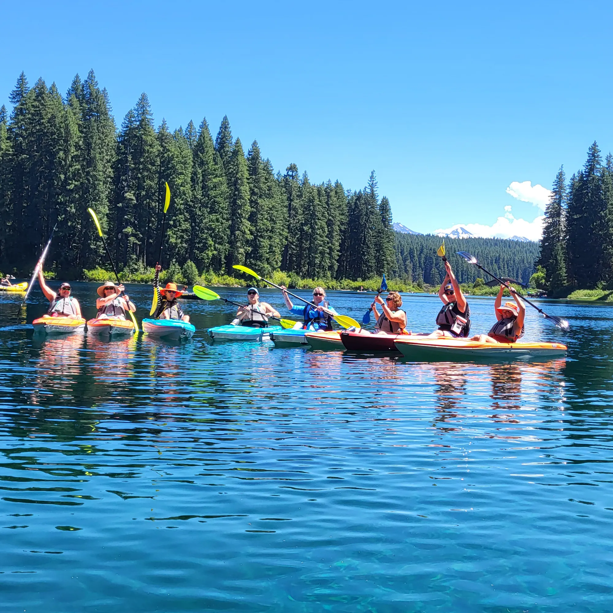 A group of people kayaking on a clear lake surrounded by lush trees and mountains under a sunny blue sky.
