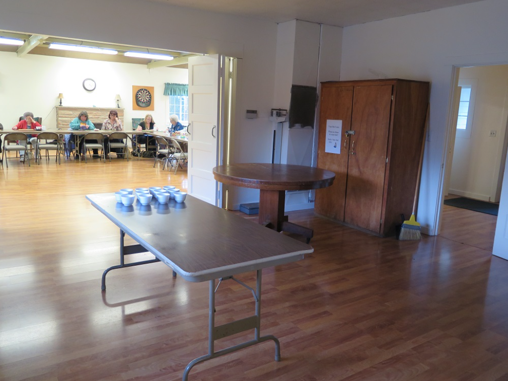 A room with people sitting at a table, a dartboard on the wall, and another table with bowls in the foreground.