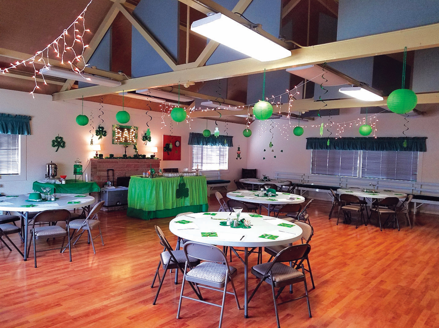Room with tables and chairs, decorated in green for a St. Patrick's Day celebration. Green lanterns and shamrocks hang from ceiling.