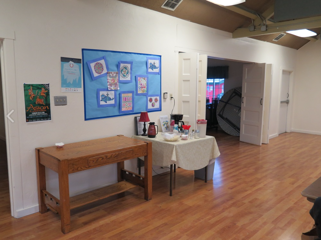 A room with wooden floors, a bulletin board, a table with coffee supplies, and a folded table by open doors.