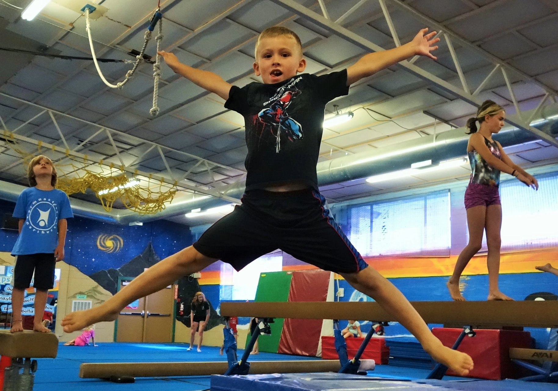 A boy balancing on a gymnastics beam with others training in the background.