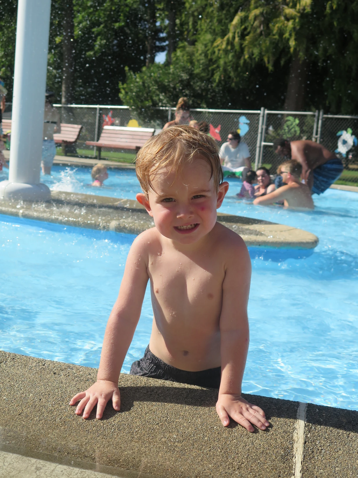 A young child at a poolside, with others enjoying water in the background.