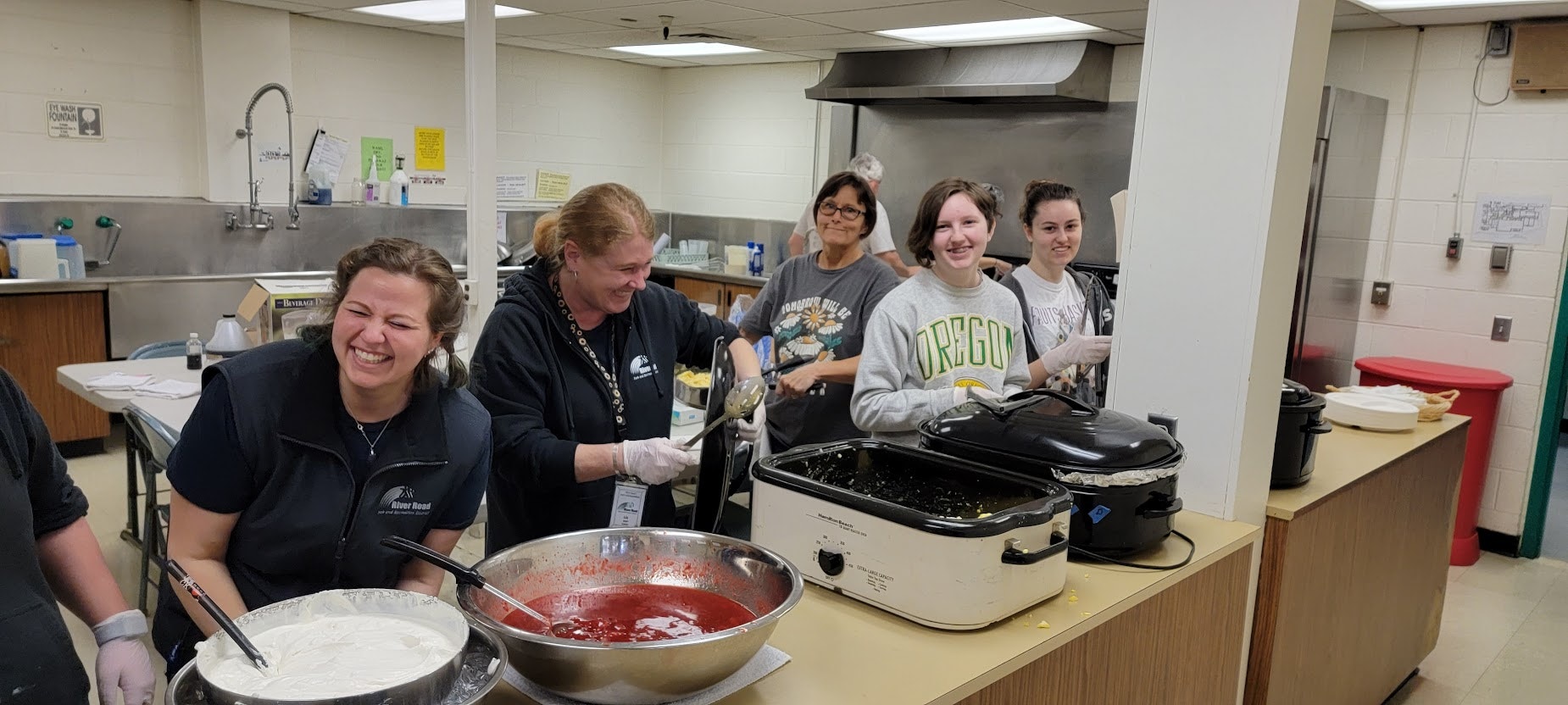 People serving food in a kitchen with big smiles.