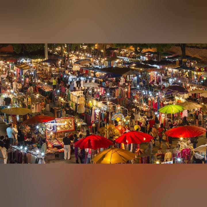 A vibrant night market filled with colorful stalls, umbrellas, and shoppers exploring various goods under twinkling lights.