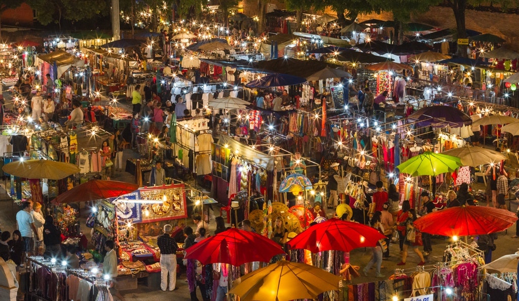 A vibrant night market filled with colorful stalls, umbrellas, and shoppers exploring various goods under twinkling lights.