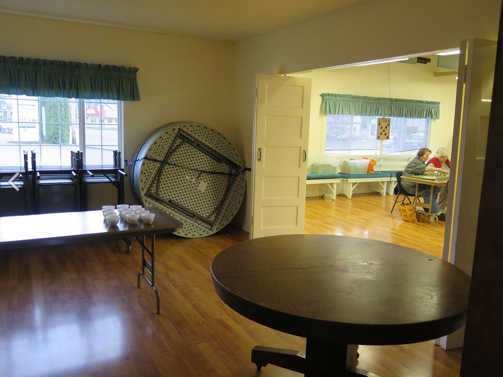 A room with tables, chairs, and a few cups. Two people sit at a table in the background.