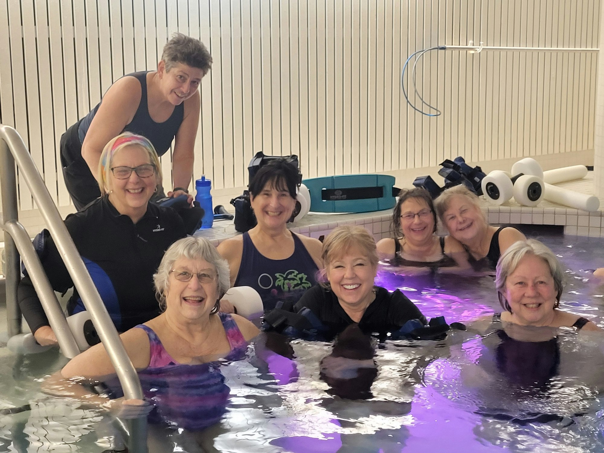 A group of eight smiling people enjoying a pool, some wearing swimming gear.