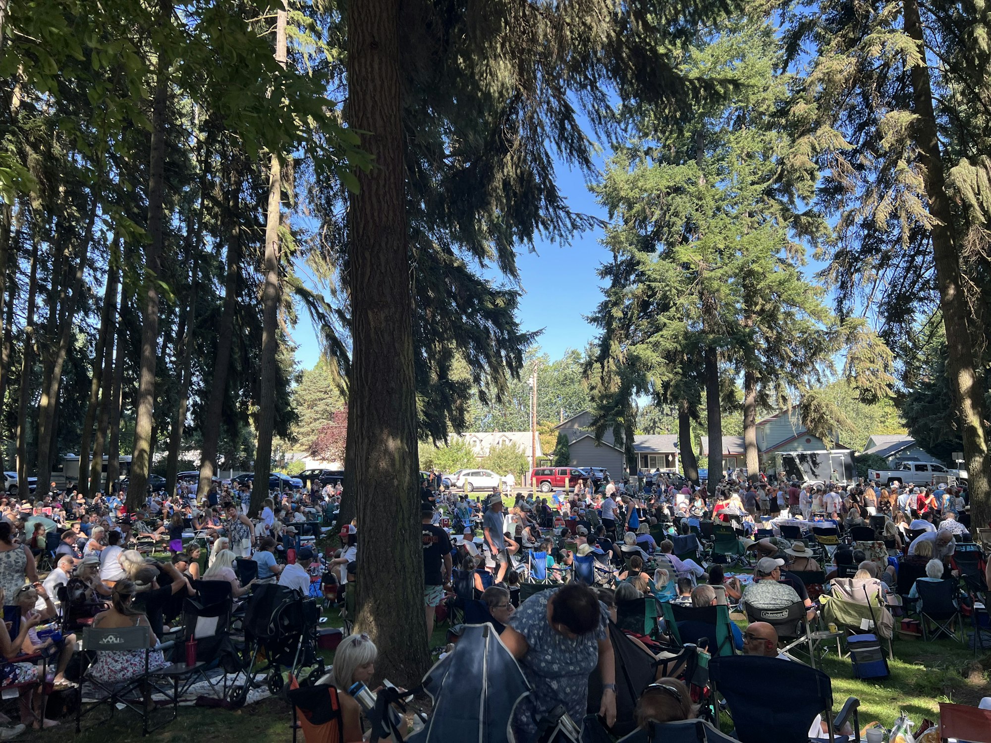 A large outdoor gathering in a wooded area with many people sitting on chairs and blankets, with houses and vehicles in the background.