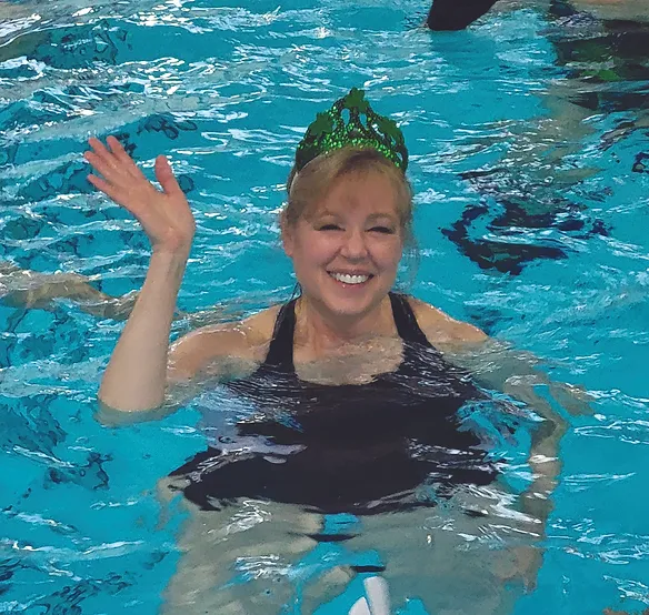 A smiling woman wearing a crown in a swimming pool, waving.