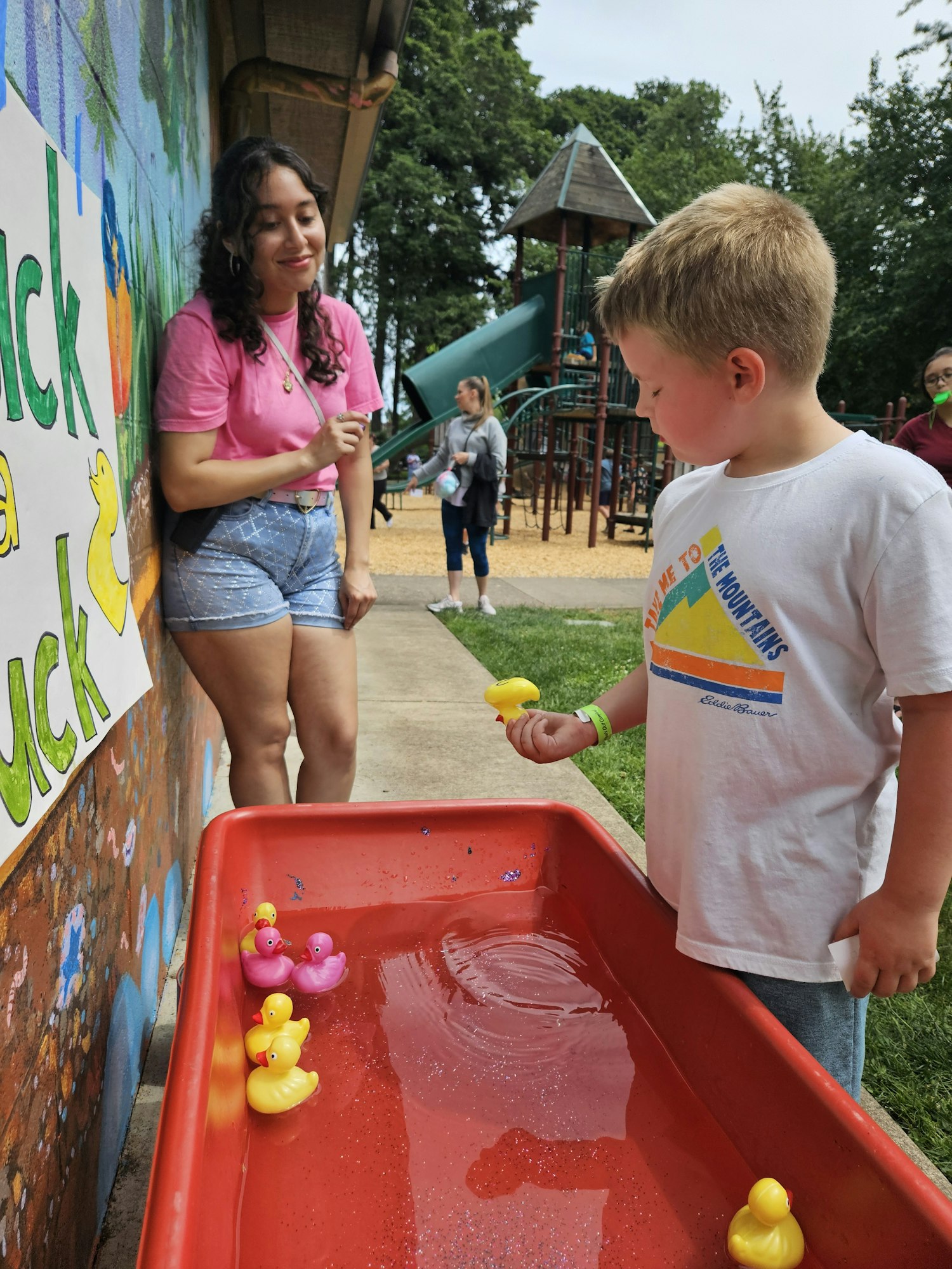 A child plays with yellow and pink rubber ducks in a water tray while a person watches, at a playground.