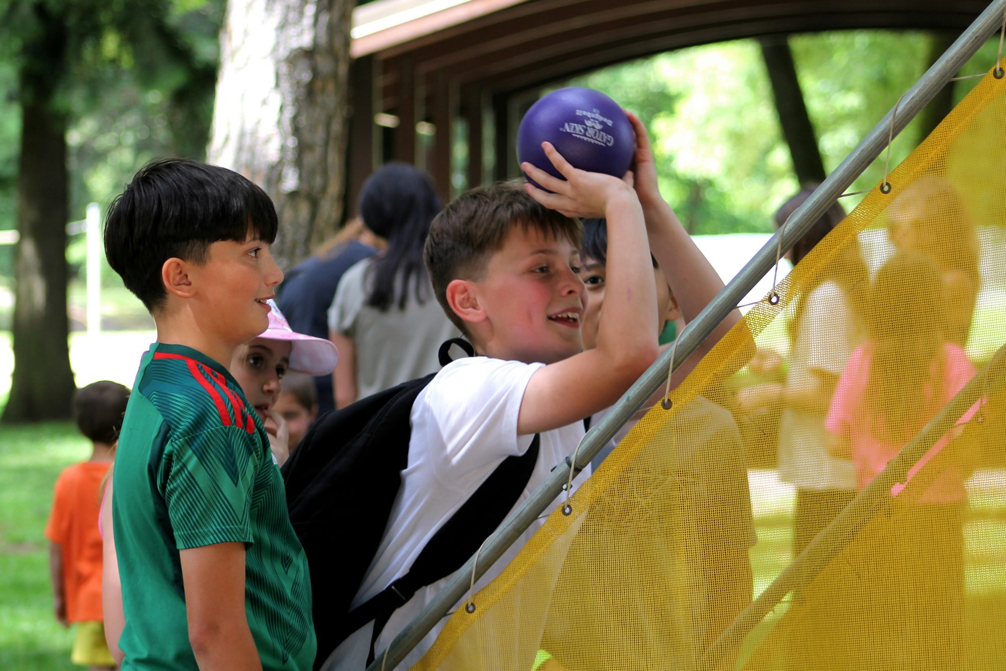 Children playing, one with a ball, outdoors near a yellow net.