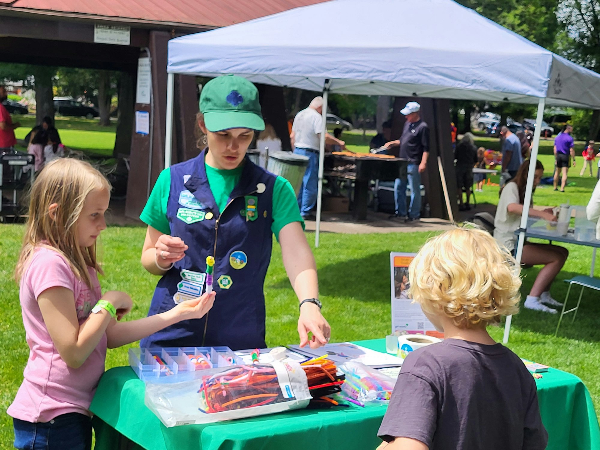 Children at a park event engage with a woman at a booth displaying crafts under a tent.