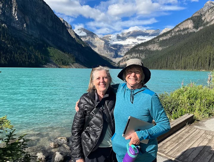 Two people smiling in front of a stunning turquoise lake with mountainous backdrop.