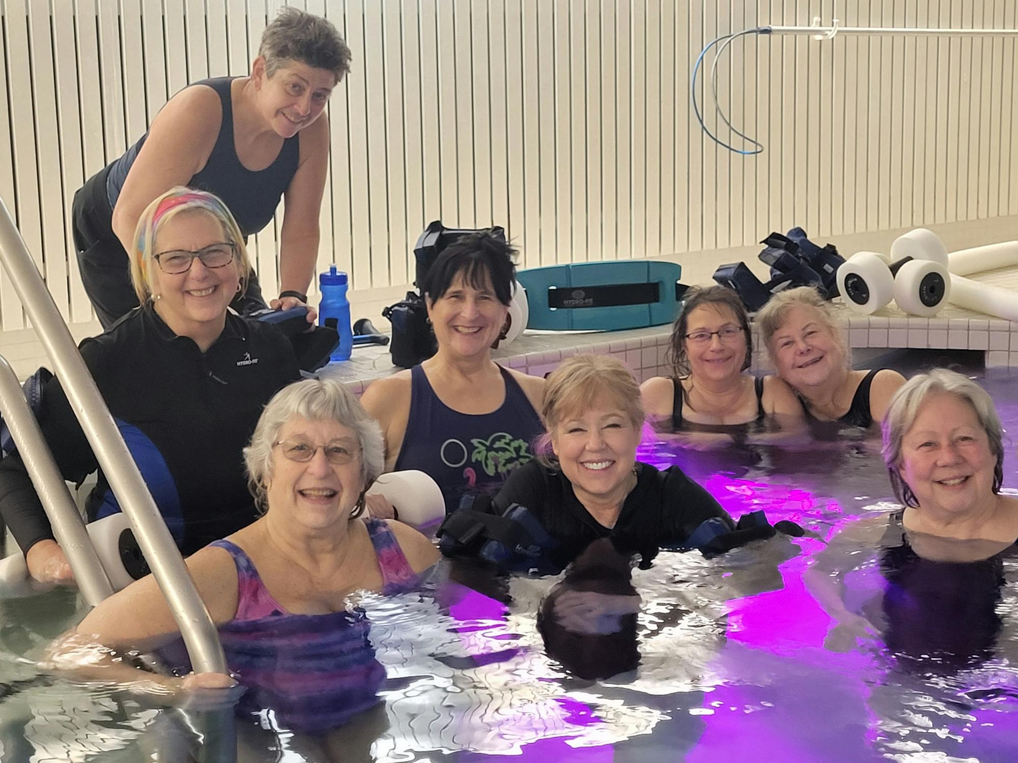 A group of women smiling while relaxing in a pool with pool equipment around them.