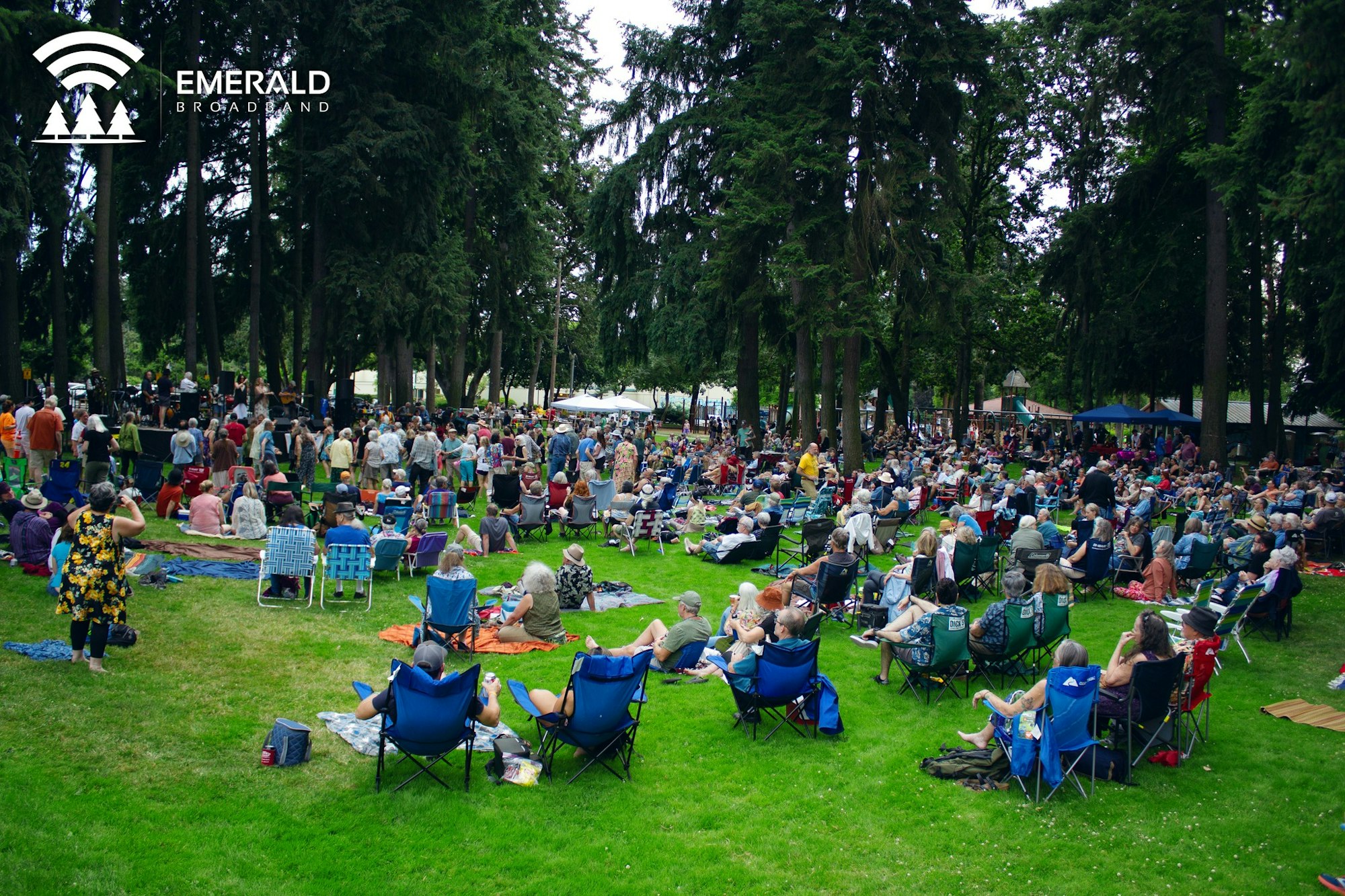 A large crowd gathered in a park with chairs and blankets, enjoying an outdoor event amid trees.