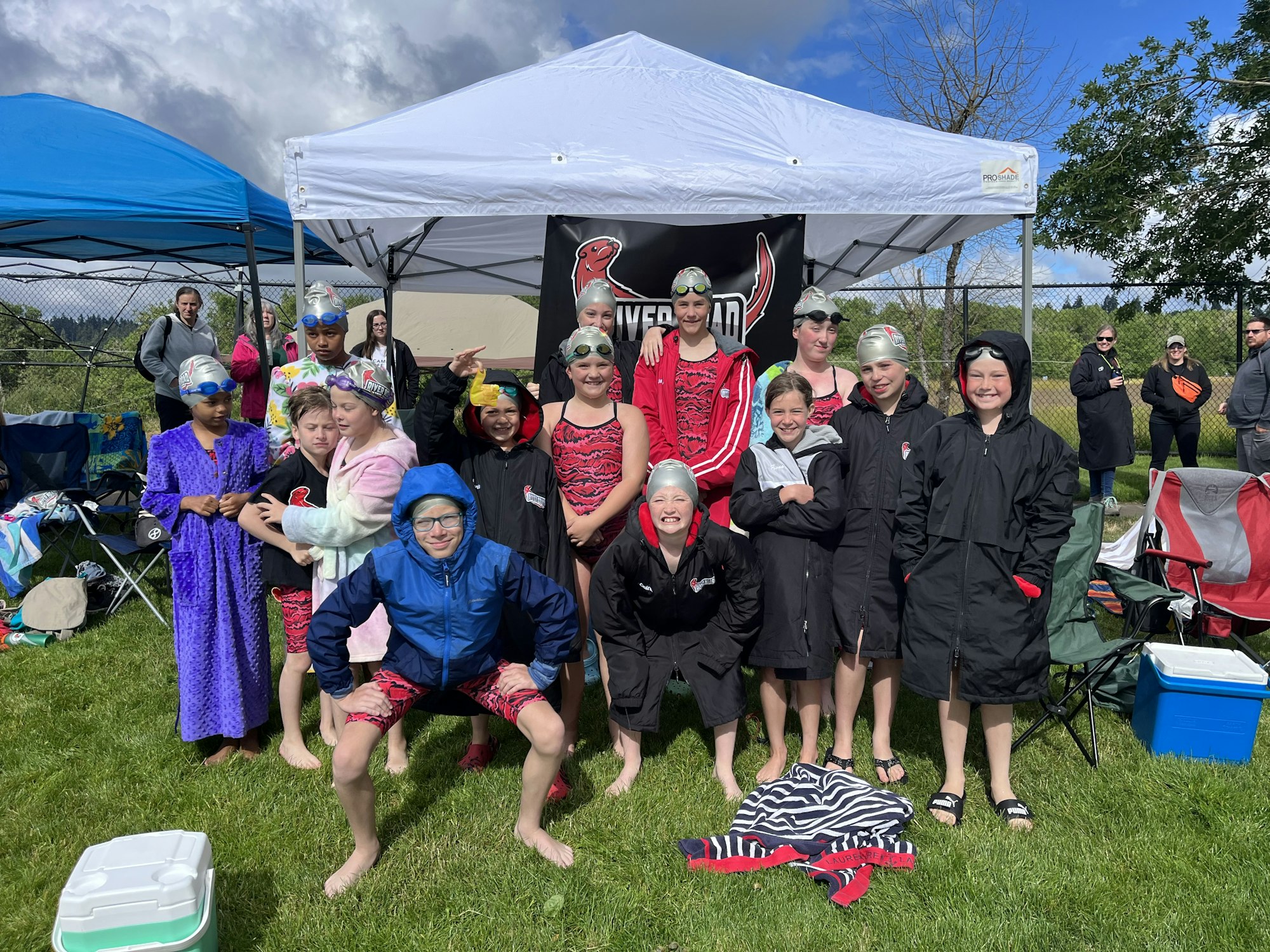 A group of swimmers in a mix of swim caps and warm clothes posing at an outdoor event, with tents and chairs in the background.