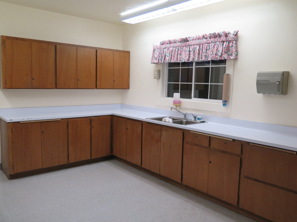 A kitchen with wooden cabinets, sink, floral curtain, paper towel dispenser, and light brown flooring.