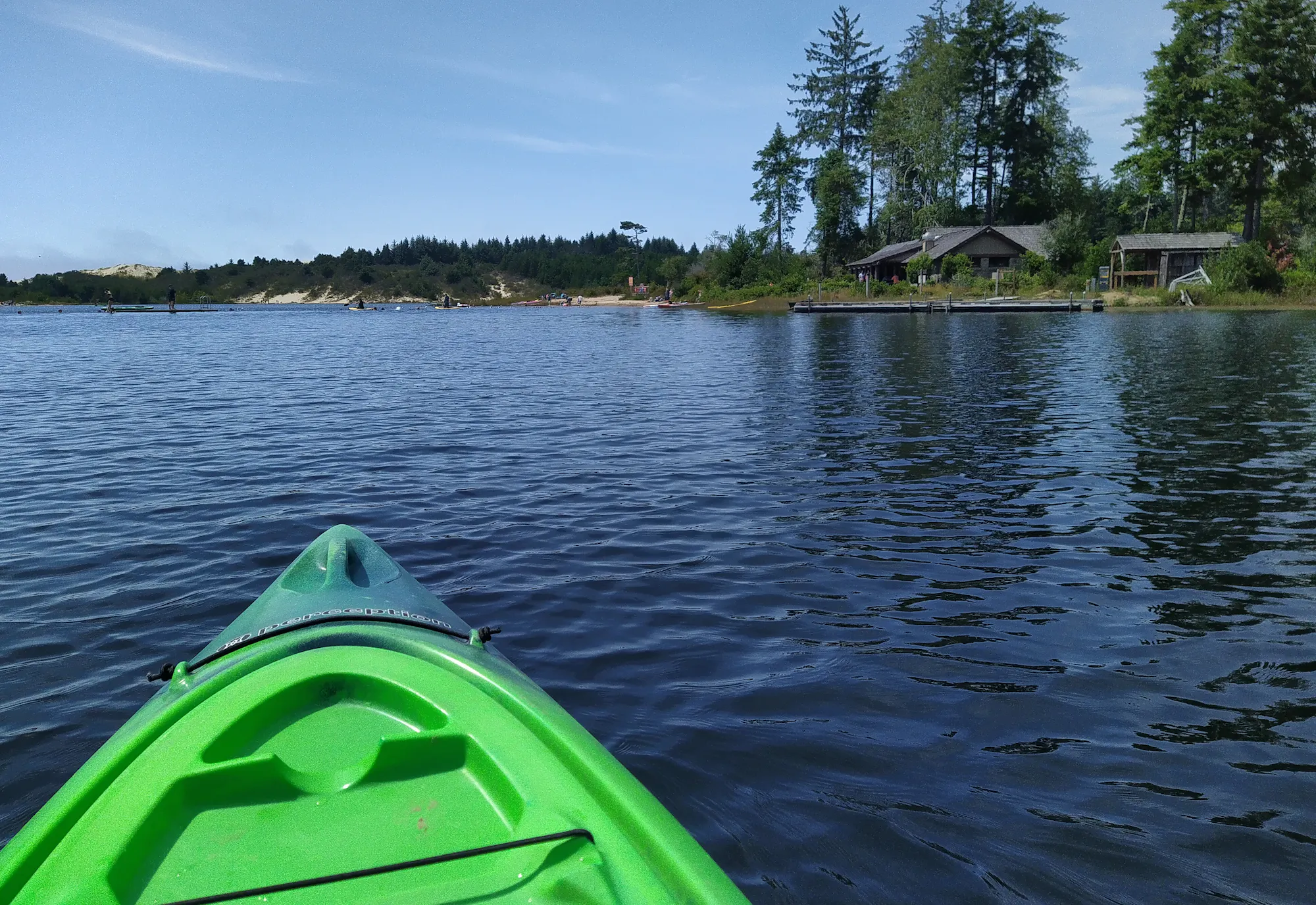 A green kayak is in calm water, overlooking a scenic shoreline with trees and houses under a clear blue sky.