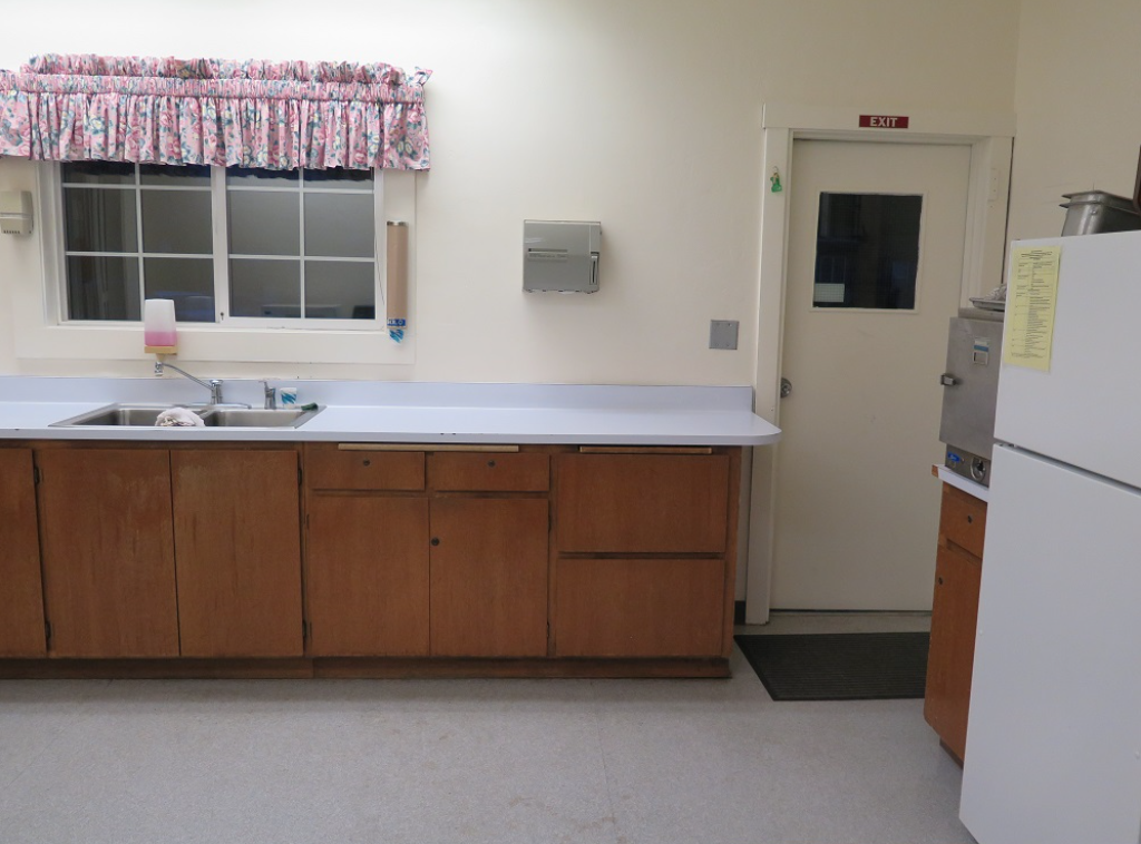 A kitchen with wooden cabinets, a countertop, sink, a door labeled "EXIT", a refrigerator, and a window with floral curtains.