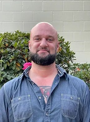 A bald man with a beard, chest tattoo, and denim shirt standing in front of a bush with a flower behind his ear.