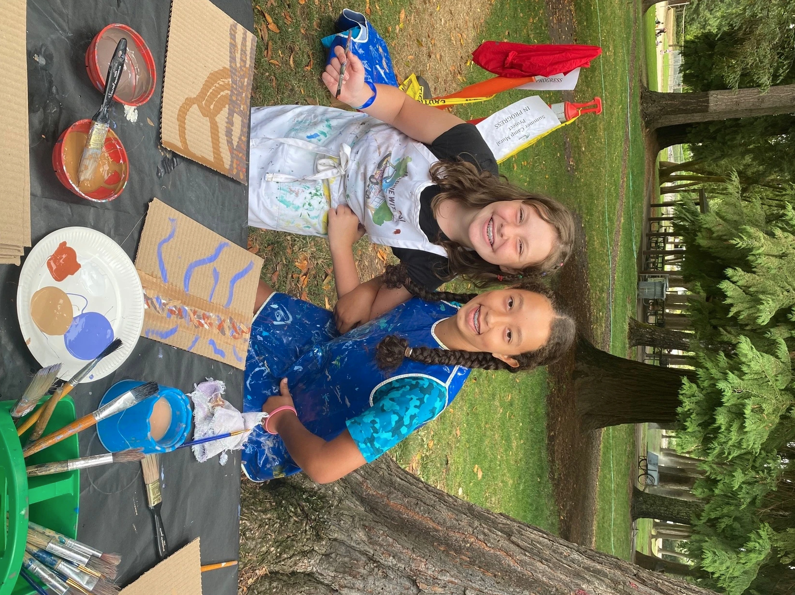 Two girls painting outdoors, with brushes, paint, and cardboard canvases on a table.