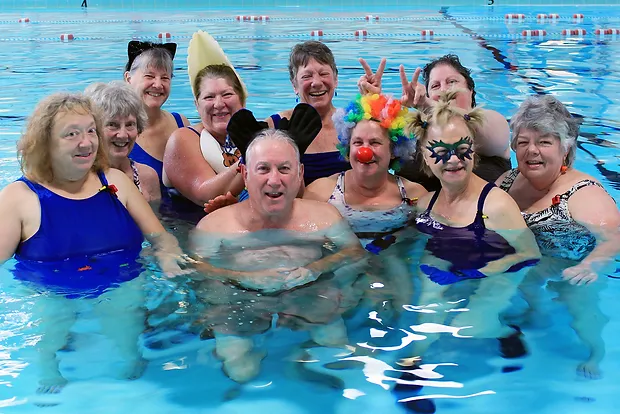A group of cheerful adults in a pool, some wearing funny hats and costume accessories.
