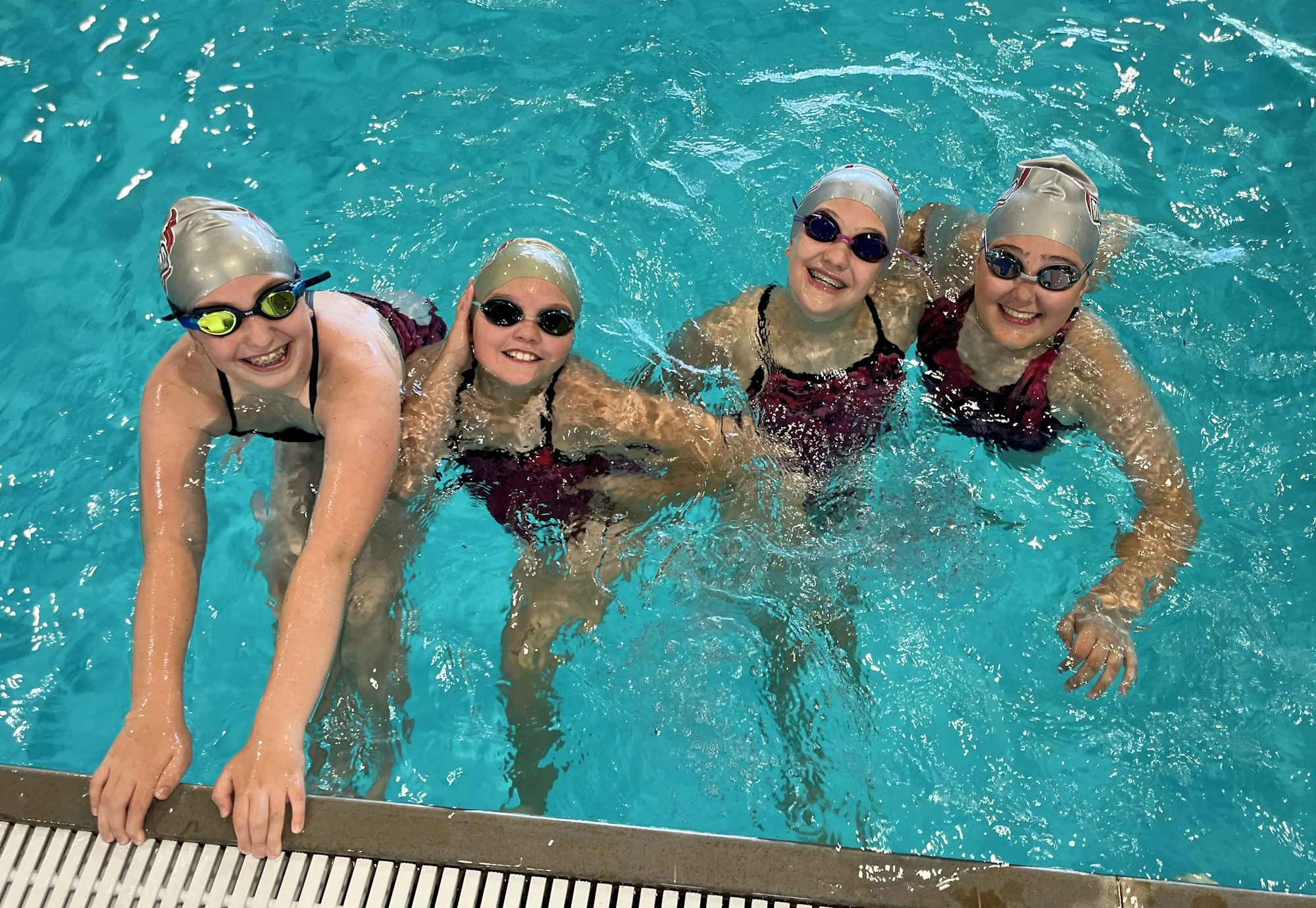 Four happy swimmers in caps and goggles at the poolside.