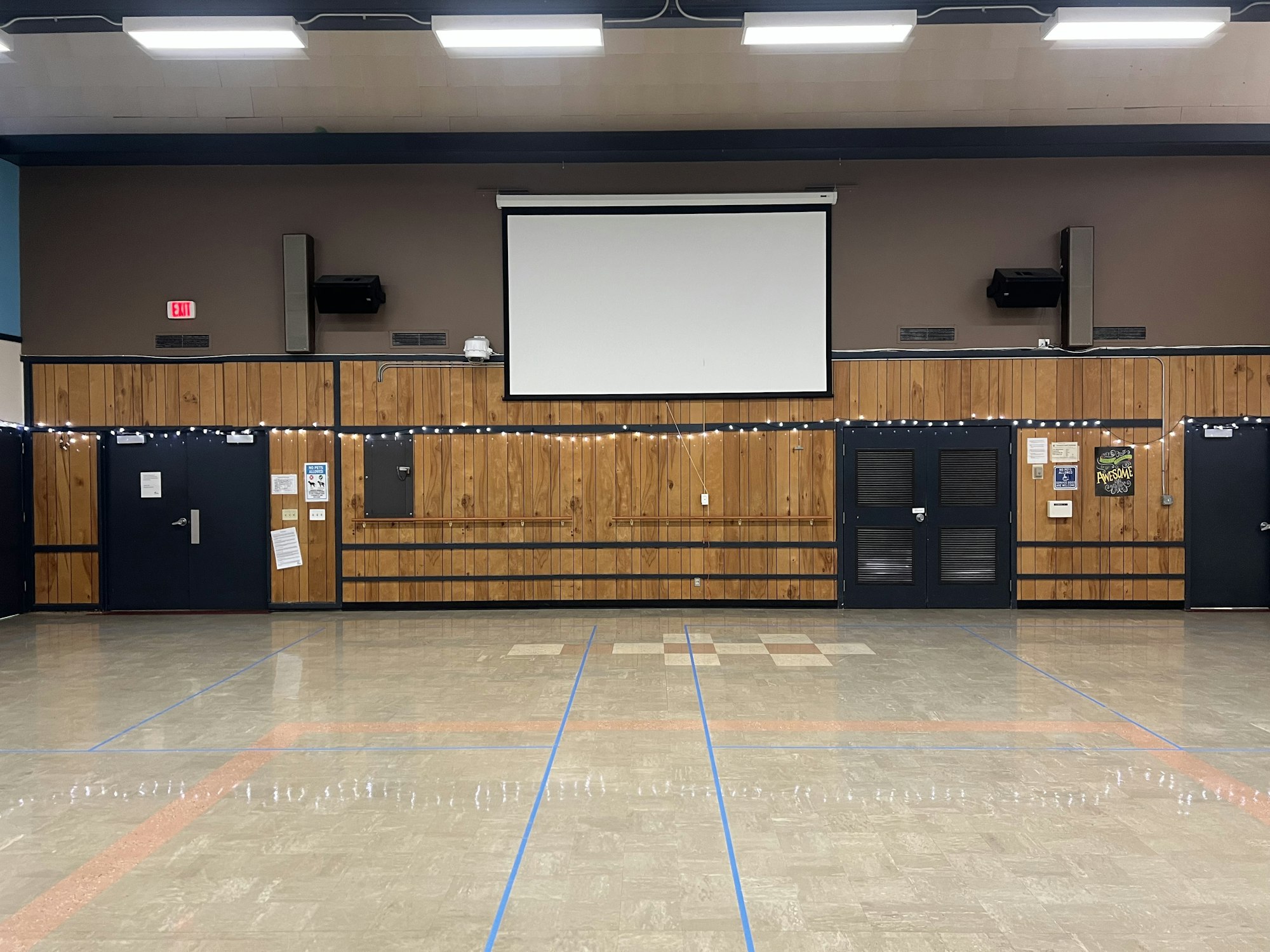 Large room with wooden panels, a projection screen, string lights, and blue doors.