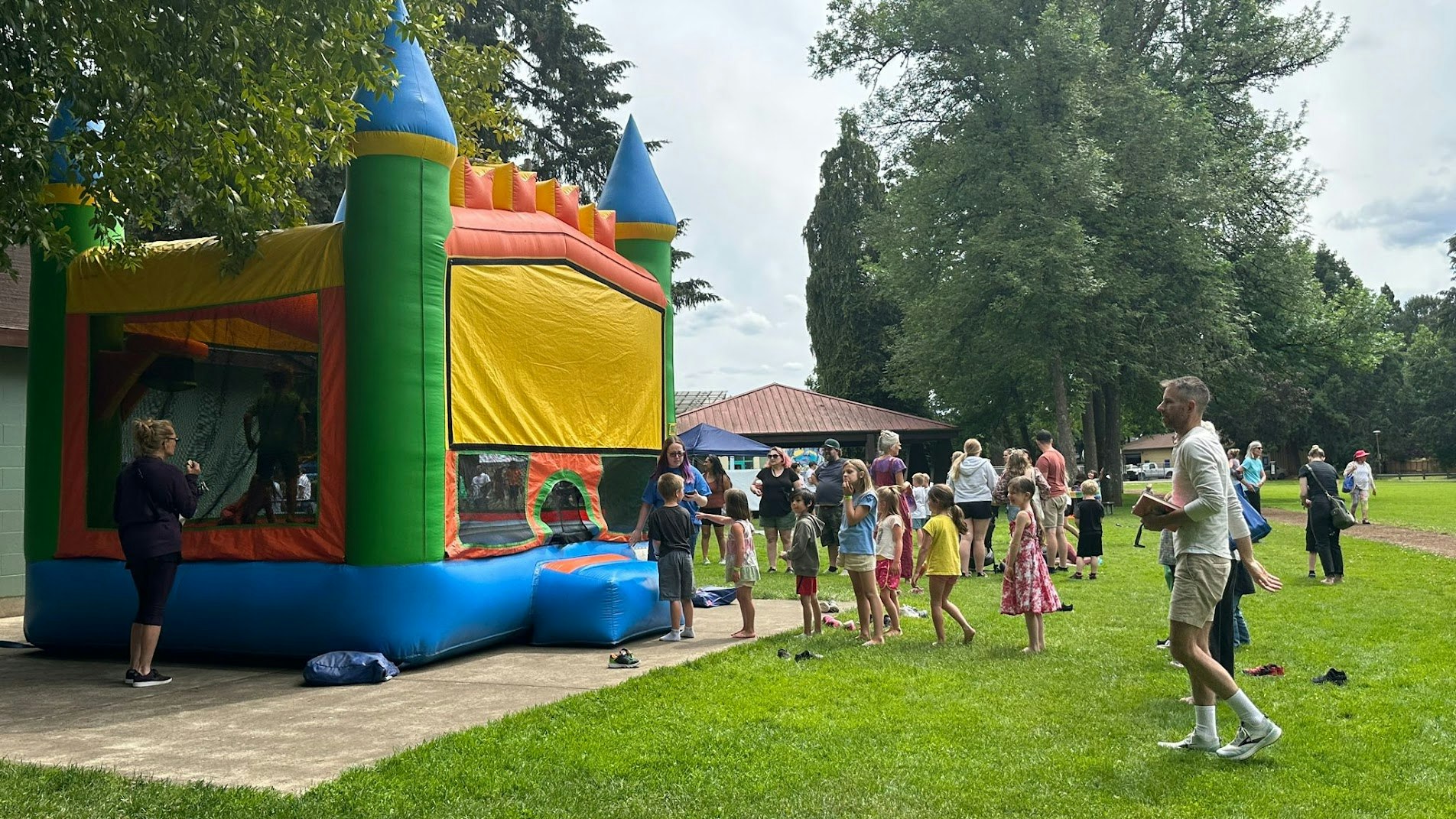 Children and adults gather around a colorful bounce house on a grassy area in a park.