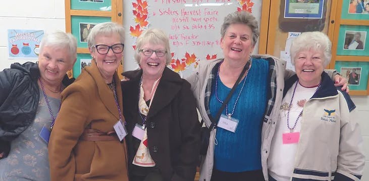 Five smiling women posing together indoors, with a cozy backdrop of fall-themed decorations and notices.