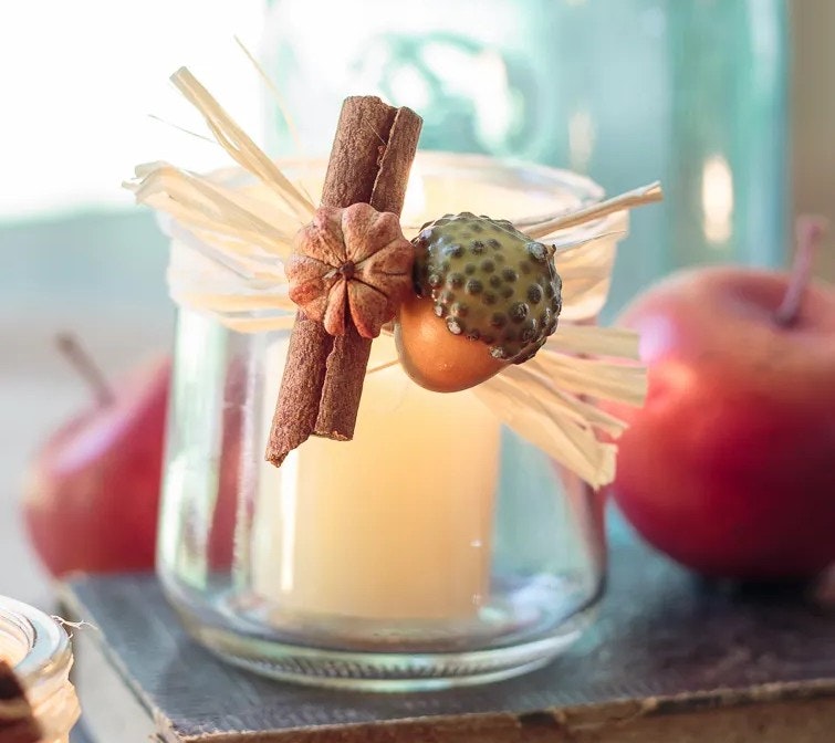A decorated candle in a glass jar with cinnamon, acorn, and straw, surrounded by apples.