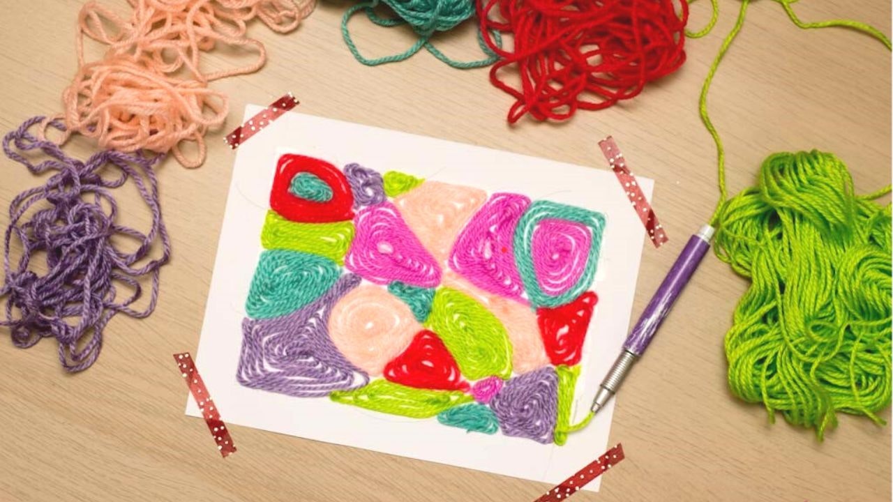 Colorful yarn art on paper with scattered yarn balls and a tool on a wooden surface.