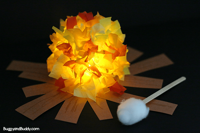 A craft project resembling a campfire made with tissue paper flame and paper logs, accompanied by a cotton swab.