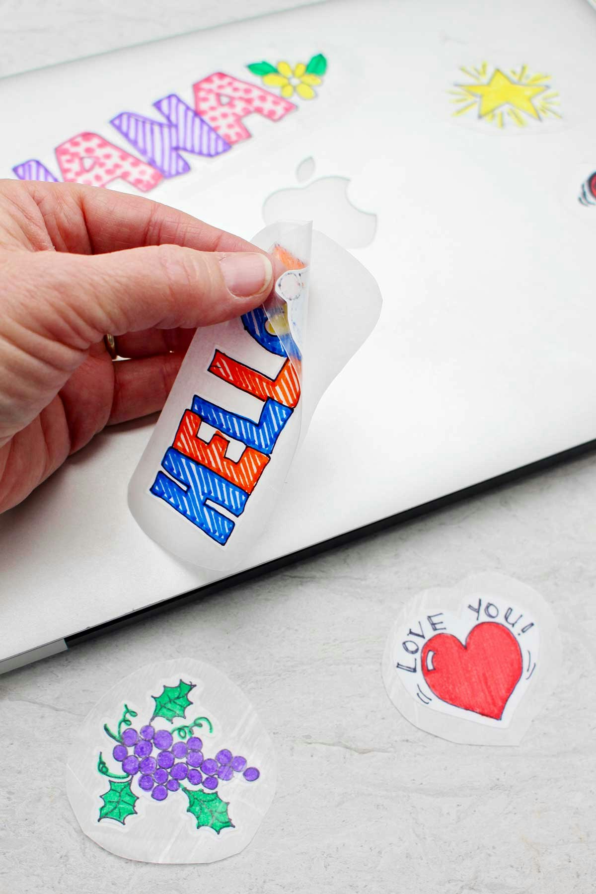 Hand peeling off a homemade sticker from a sheet placed on a laptop, with more colorful stickers around.