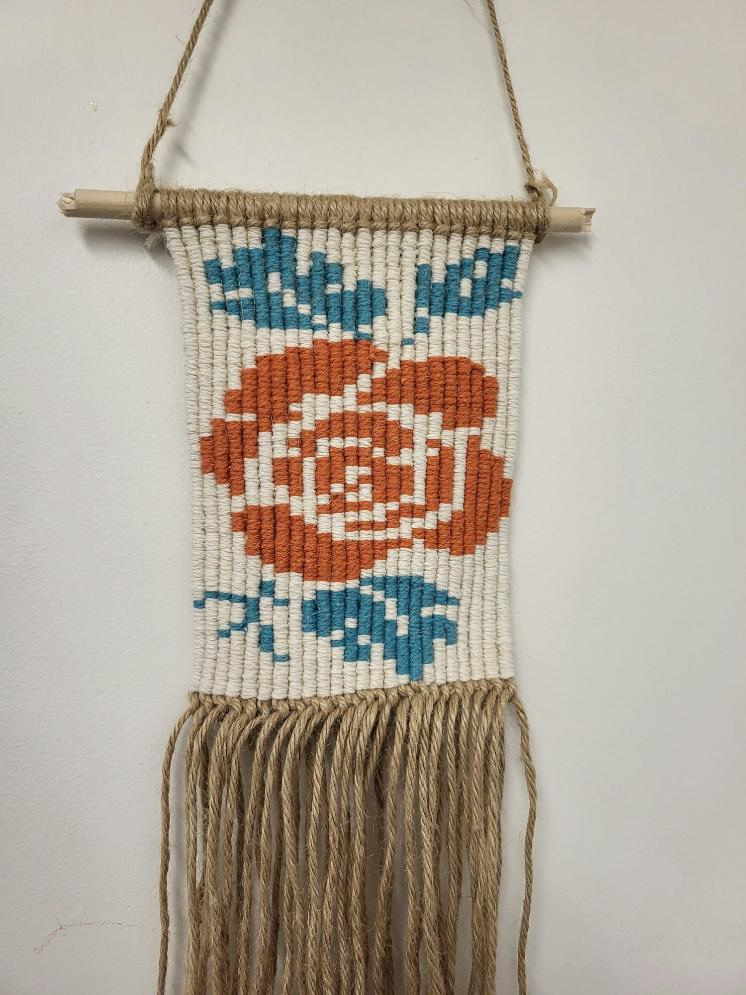 Woven wall hanging with a red flower and blue leaves, with fringe on the side.
