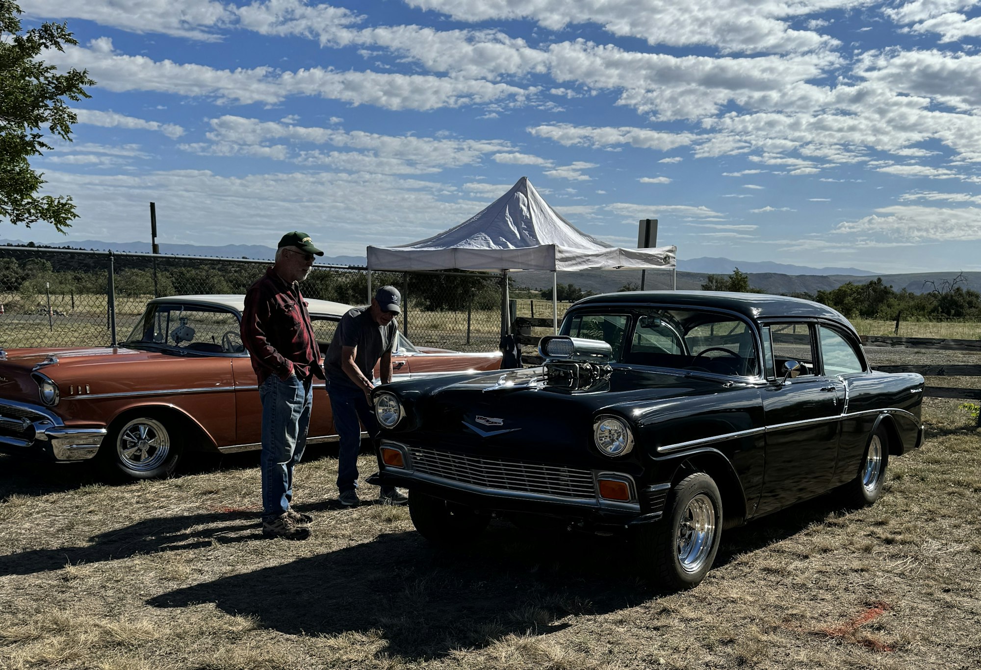Two men with vintage cars, one with an open hood, outdoors under a cloudy sky.