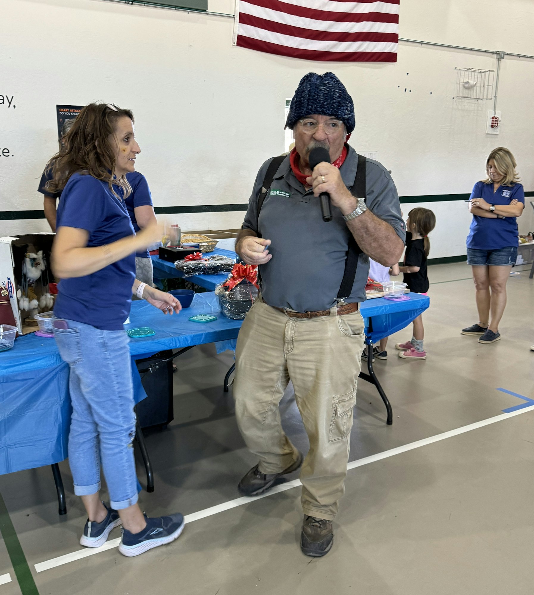 A man in a hat speaks into a microphone at an indoor event with tables and a US flag in the background.