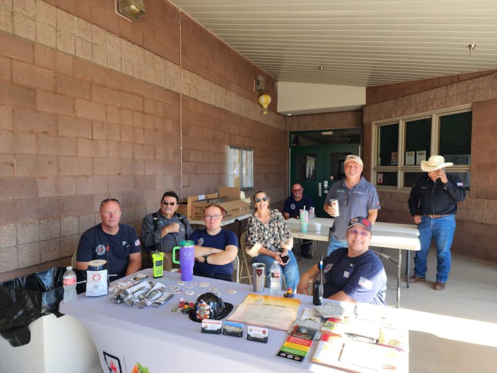 Group of people sitting and standing around tables with drinks and pamphlets outside a brick building.