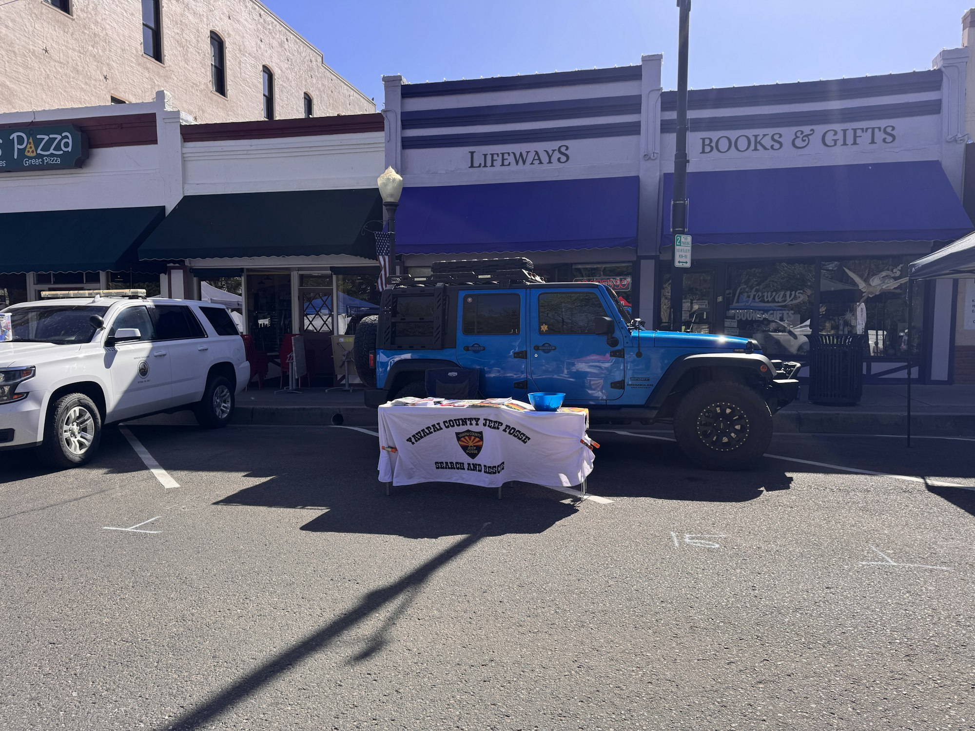 A blue Jeep and a white SUV parked outside shops named "Lifeways" and "Pazza," with a table for a local search and rescue team.