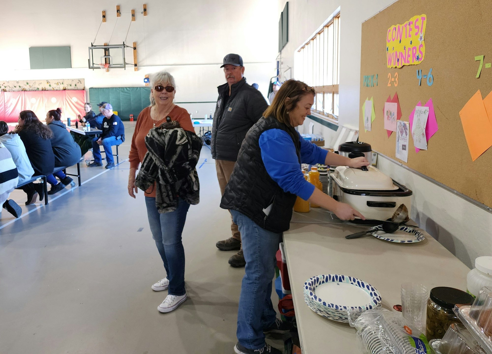 Indoor community event with people serving food, chatting, and a contest winners board in the background.