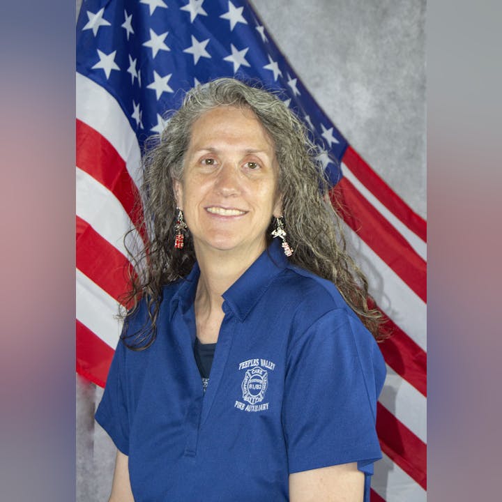 A woman in a blue shirt with "Peeples Valley Fire Auxiliary" embroidered on it, smiles before an American flag.