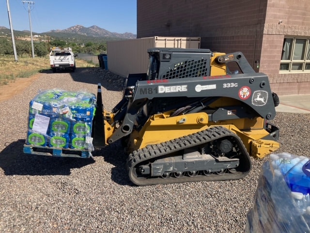 A skid steer loader transporting a pallet of bottled water, with a police vehicle and mountainous landscape in the background.