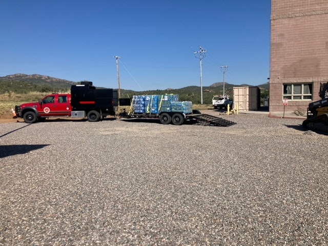 Truck with flatbed trailer carrying water bottles parked outside a building, with clear sky and hills in the background.