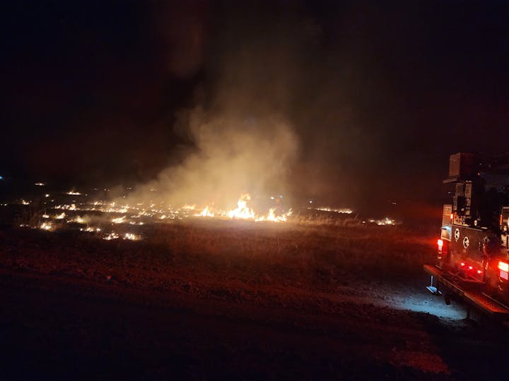 A nighttime scene of a grassland fire with flames and smoke, alongside a fire truck in the foreground.