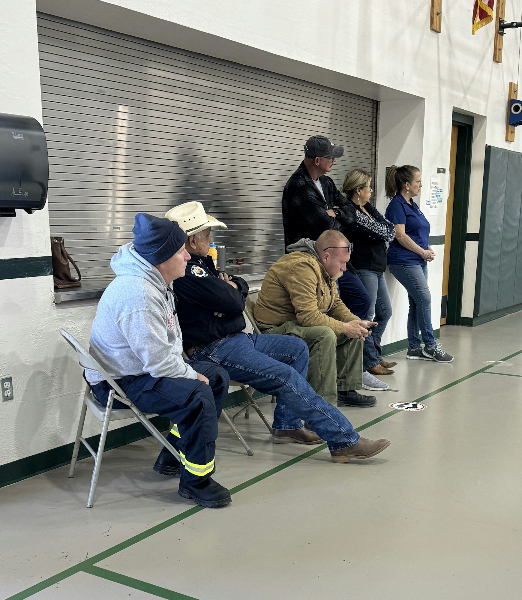 People sitting and standing in a room, some appear to be casually gathered, others are seated on folding chairs, next to a wall.
