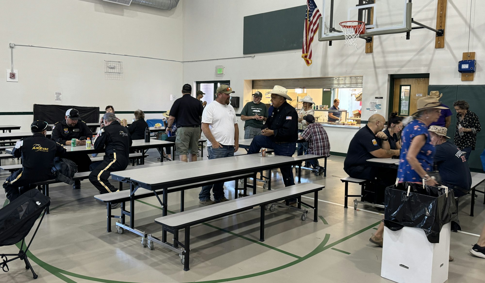 People socializing in a gymnasium with tables, a basketball hoop, and an American flag.