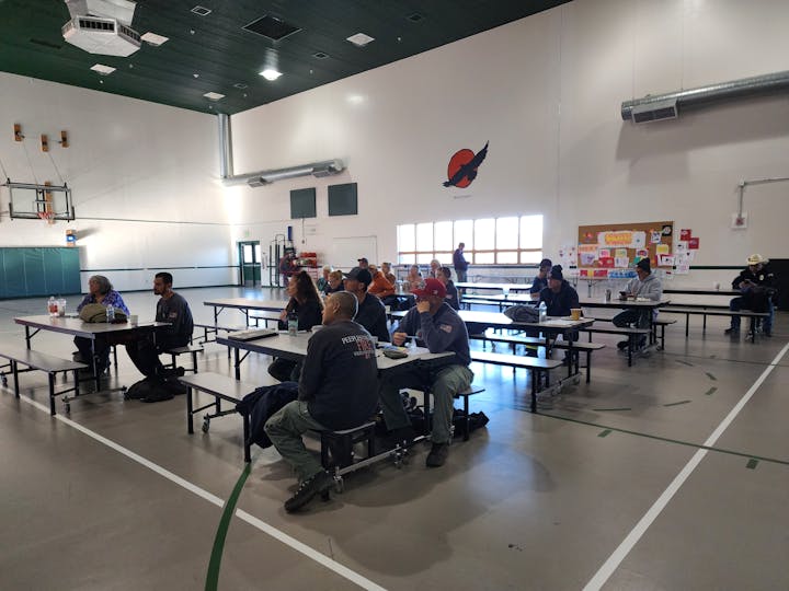 A group of people sitting at tables in a gymnasium, some eating, with a basketball hoop and colorful bulletin board in the background.