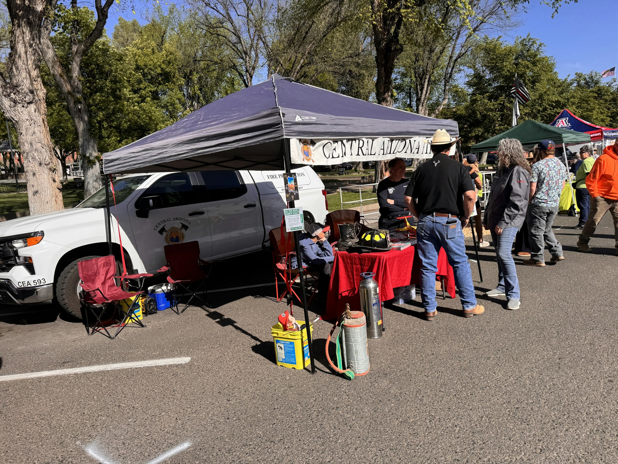A booth at an outdoor event featuring Central Arizona fire prevention displays, people mingling, and a parked vehicle.