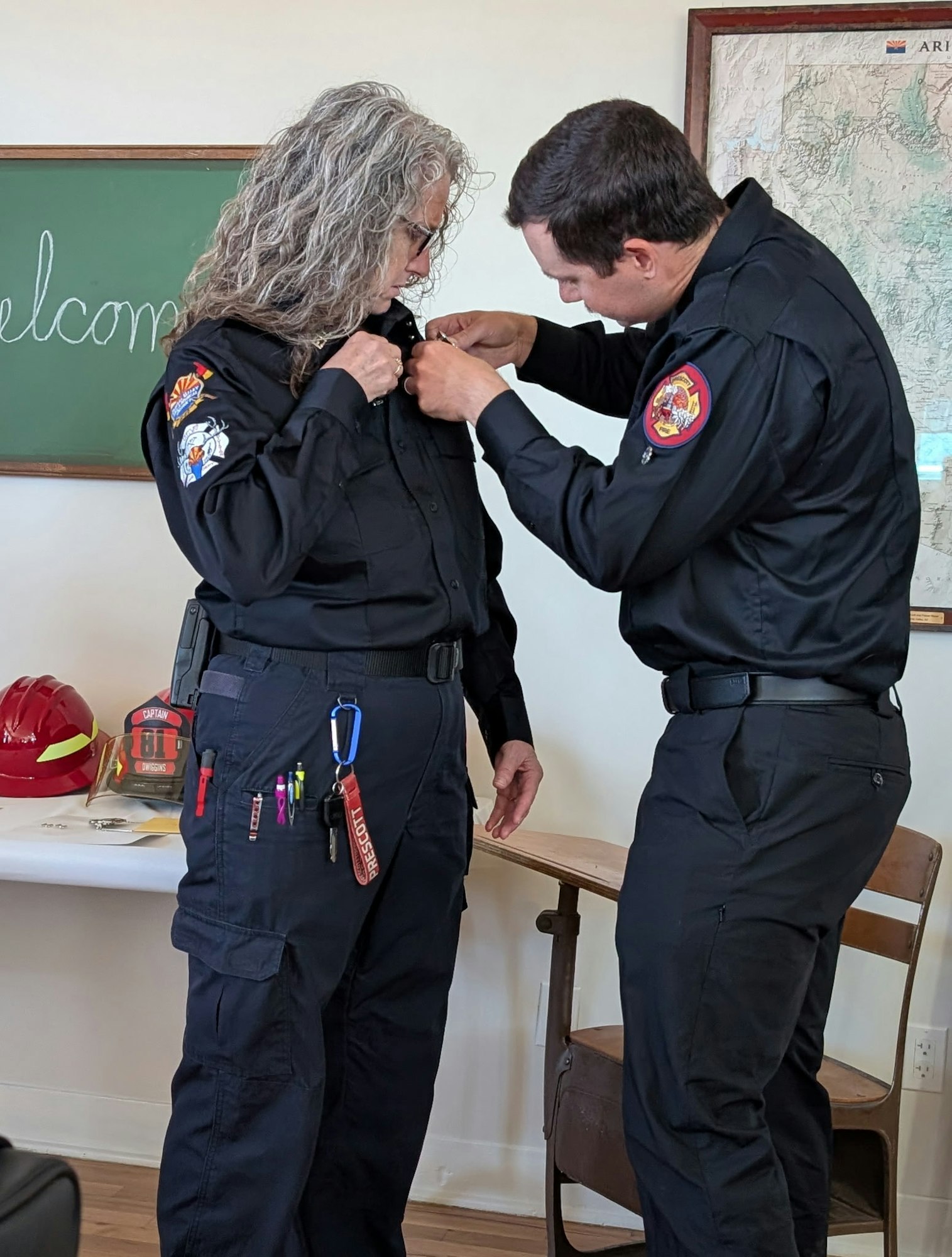 A woman in a uniform is being assisted by a man to adjust her shirt, with a classroom setting and fire gear in the background.