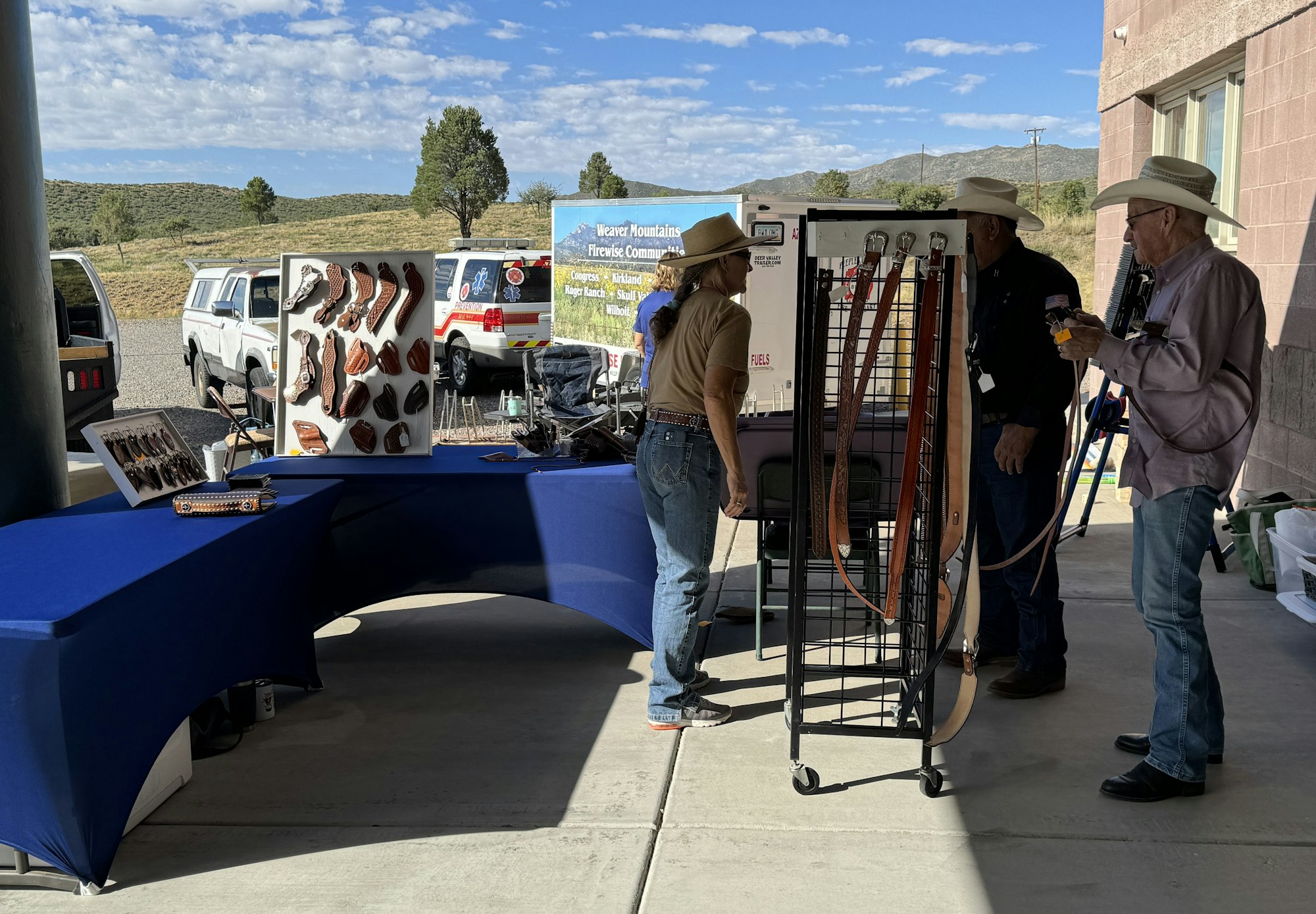 Outdoor event with leather goods on display, people in cowboy hats, ambulance in the background, sunny day.